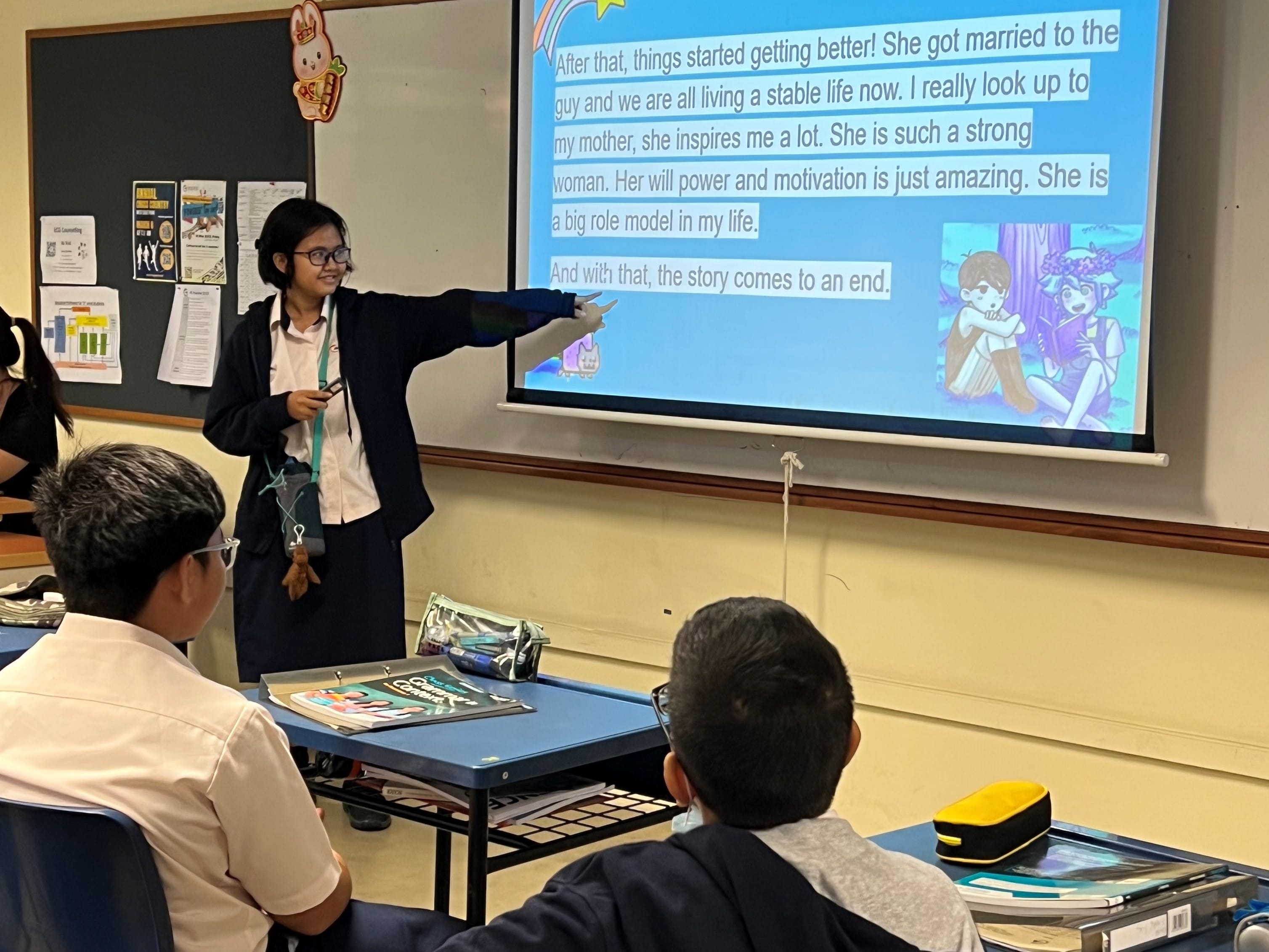 A student in a blazer points at a projected slide with text about a mother as a role model, while classmates watch.